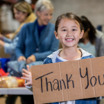 A young girl holds up a hand-written thank you sign in front of a backdrop of tables, and volunteers