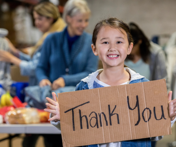 A young girl holds up a hand-written thank you sign in front of a backdrop of tables, and volunteers