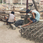 Older children at a rural Myanmar shelter watching and helping build new building support columns during summer construction.