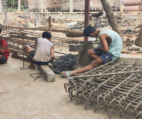 Older children at a rural Myanmar shelter watching and helping build new building support columns during summer construction.