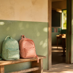 Two small children's backpacks resting side by side by a sunlit classroom doorway, representing two siblings safe and in school at a Project Nic shelter.