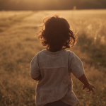 A young child runs through a sunlit open field, seen from behind, evoking freedom and safety.
